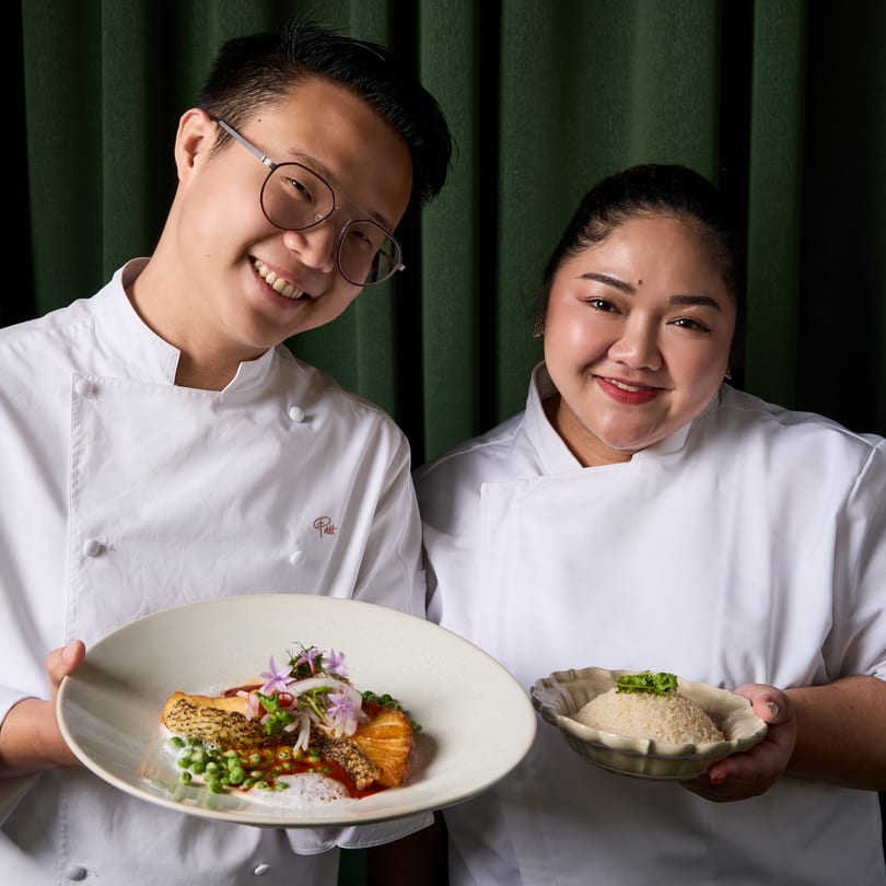 Two smiling Khaosan chefs in white coats hold a salmon dish and a bowl of rice.