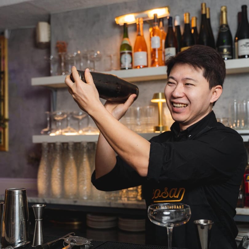A smiling bartender in a black apron shakes a cocktail shaker at Khaosan Thai restaurant.
