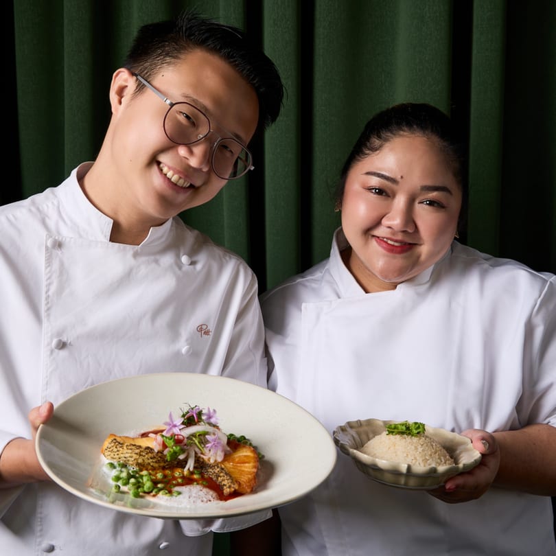 Two smiling Khaosan chefs in white uniforms holding a Thai fish dish and a bowl of rice.