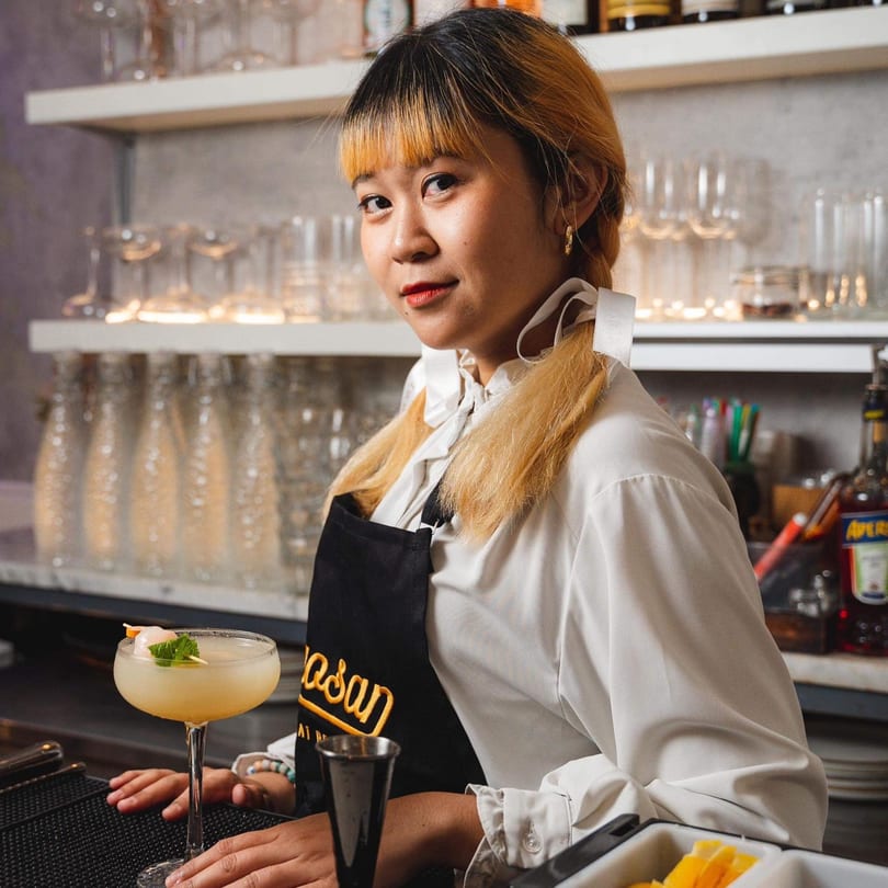 A Khaosan bartender in a white shirt and black apron, with a cocktail on the bar.