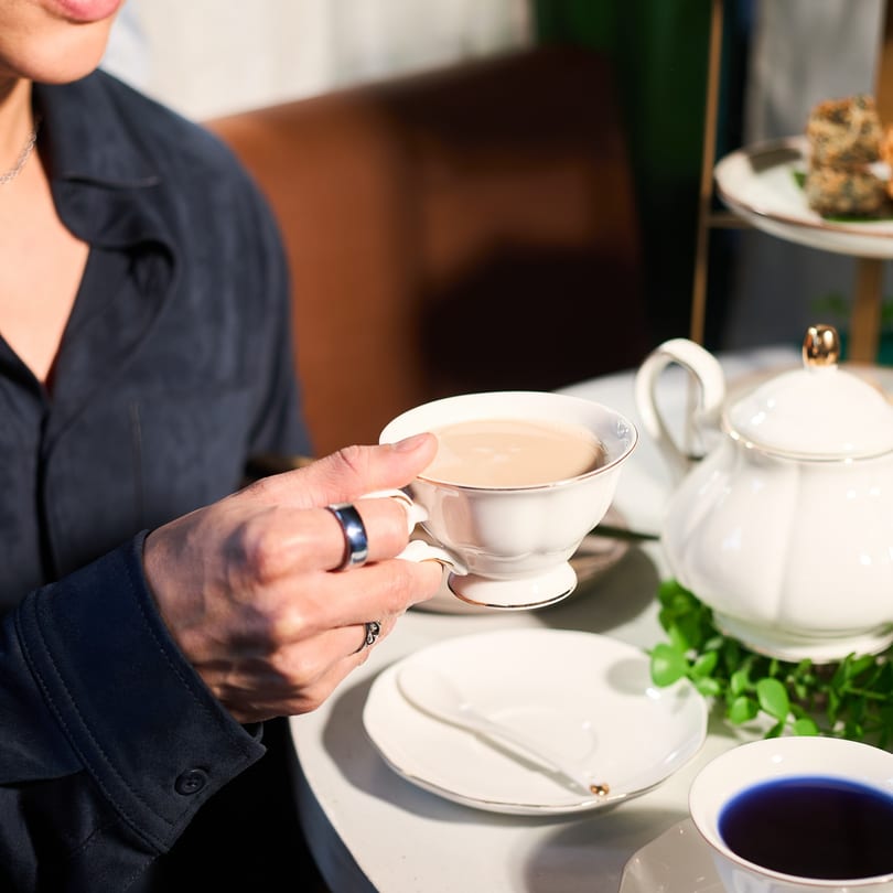 A person holding a white teacup filled with light brown Thai tea, with a teapot nearby.