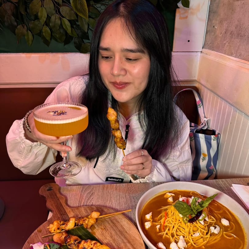Woman at Khaosan Thai restaurant holding a cocktail and chicken skewer, with a noodle dish on the table.