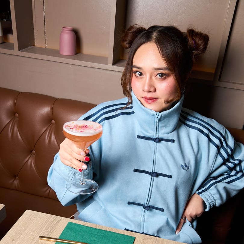 A young woman with space buns holds up a pink cocktail in a Thai restaurant.