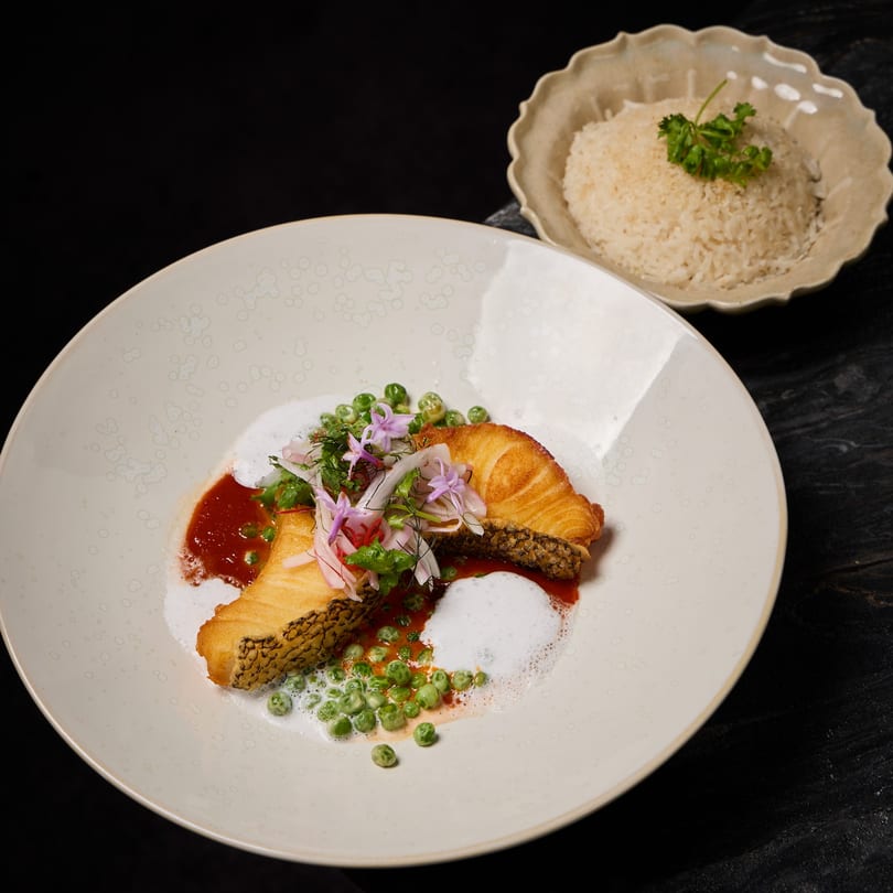 Pan-fried fish with crispy skin, red sauce, green peas, and edible flowers, served with rice.