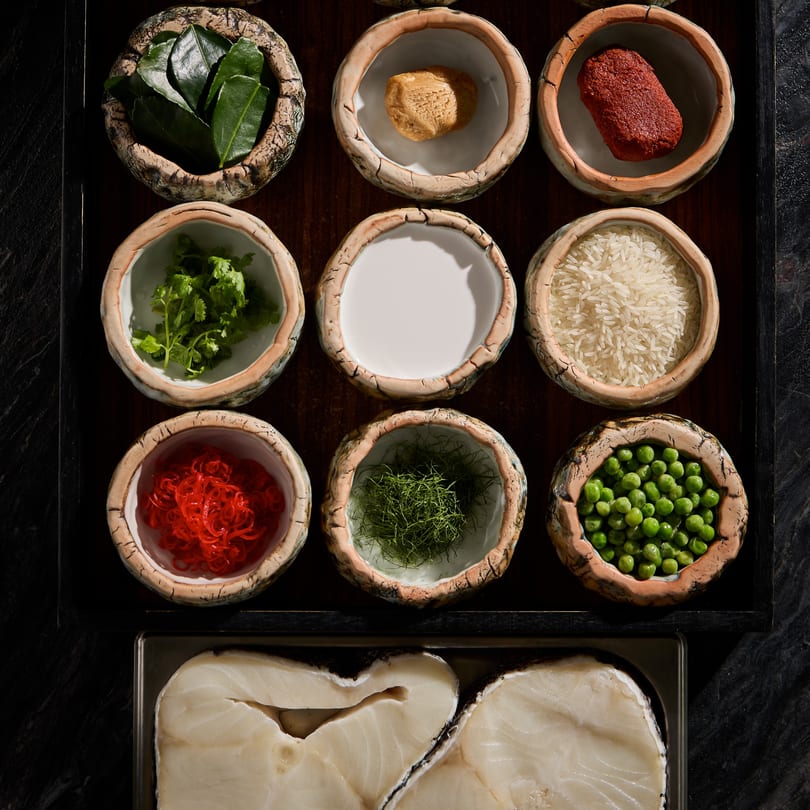Nine bowls of Thai ingredients, including herbs, spices, rice, and peas, with raw fish below.