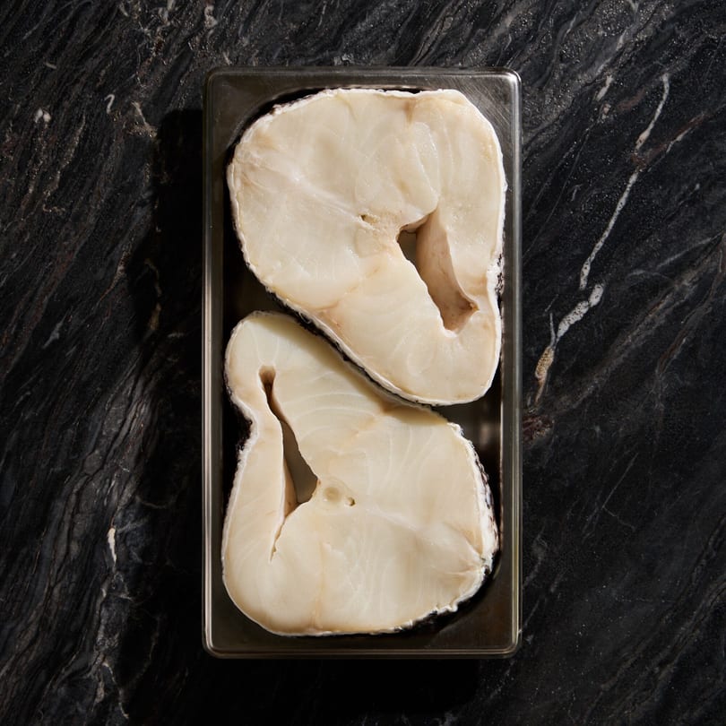 Two raw, thick-cut fish fillets in a metal tray on a dark marble surface.