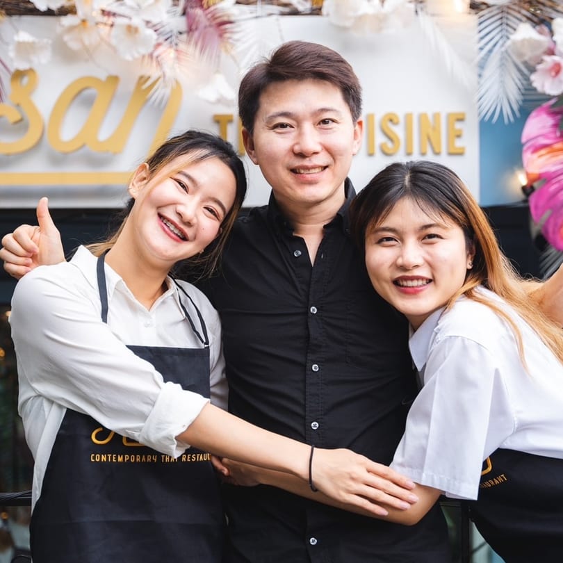 Three smiling Khaosan Thai restaurant staff members, two women and one man, posing happily together.