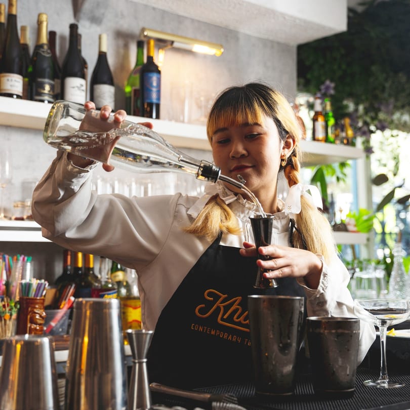 A Khaosan bartender in an apron pours liquid from a bottle into a measuring jigger.