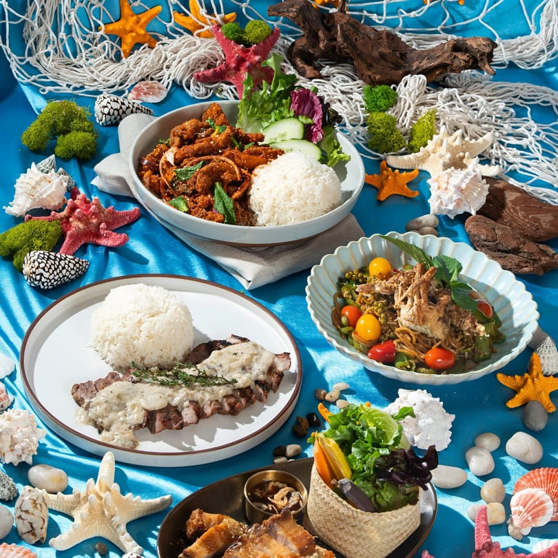 Three Thai dishes, including steak with sauce, on a blue tablecloth with seashells and starfish.