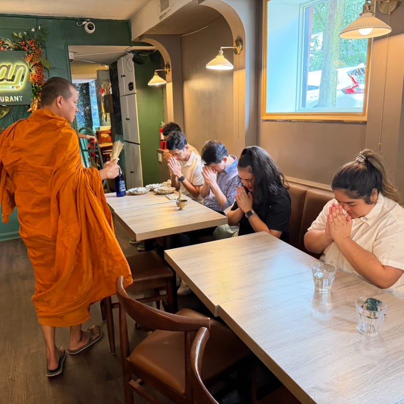 A monk in orange robes blesses people seated at tables in a Thai restaurant.