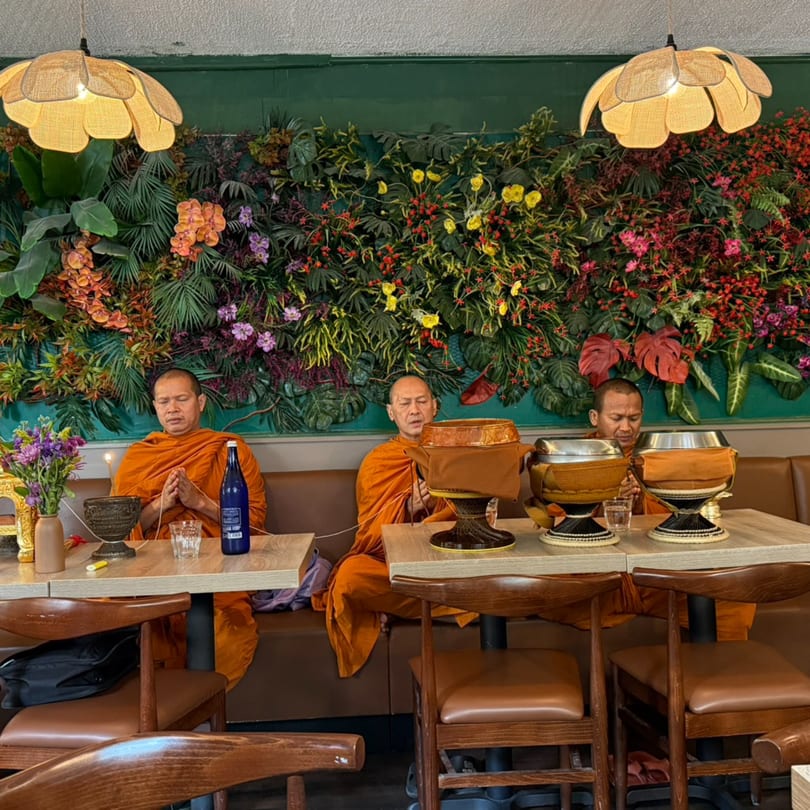 Three monks in orange robes praying at tables in a Thai restaurant with a floral wall.