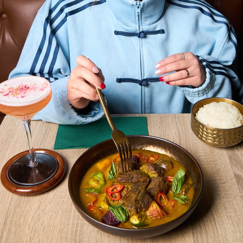 A person in a blue jacket eating Khaosan Thai green curry with rice and a cocktail.