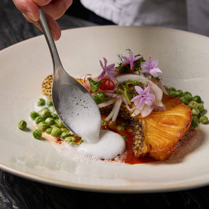 A chef garnishes a Thai fish dish with foamy sauce, peas, and edible flowers.