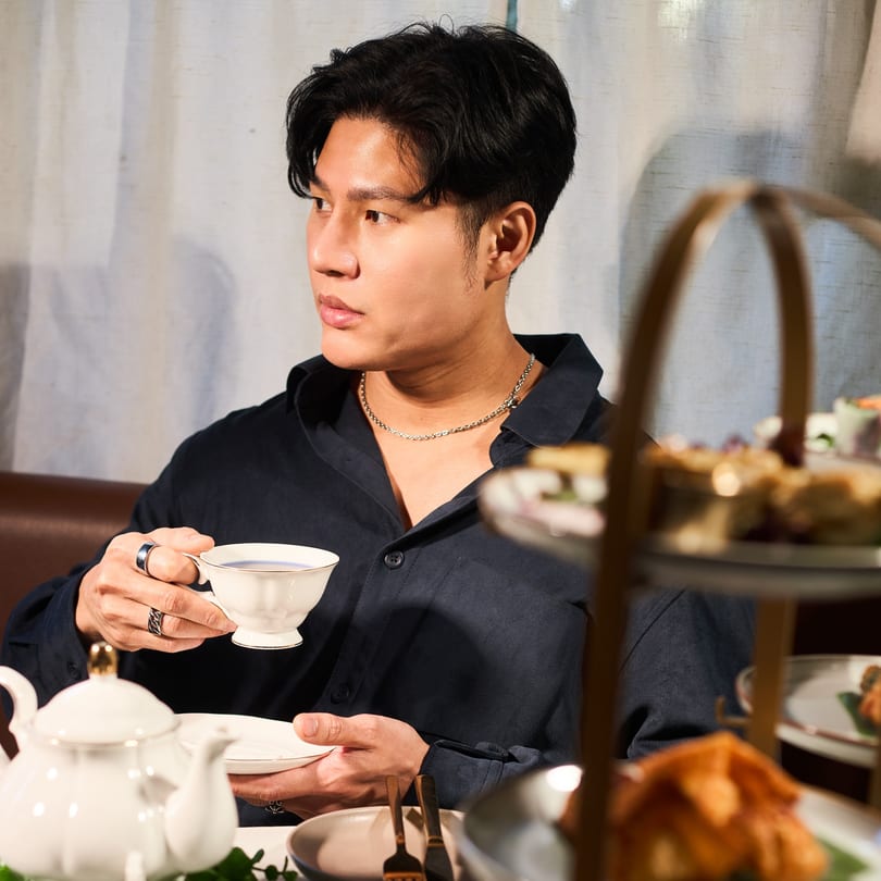 A man in a dark shirt sips tea from a white cup at a table with a teapot and tiered dessert stand.