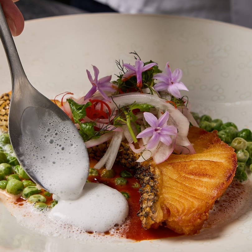 A spoon pours white foam over a Thai dish with fried fish, green peas, red sauce, and purple flowers.