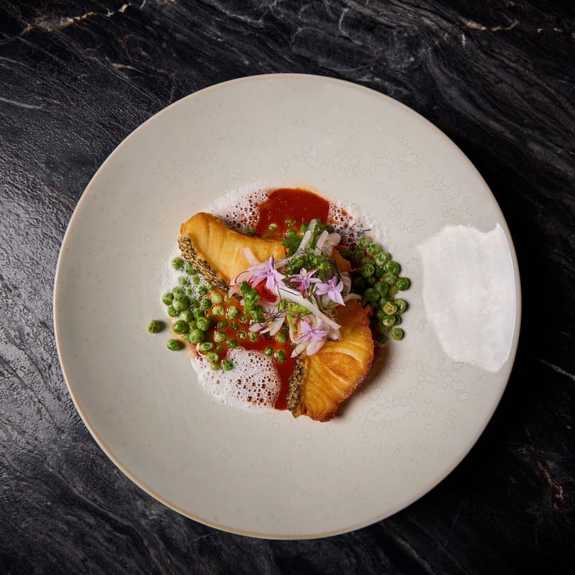 Crispy fried fish with red sauce, green peas, and delicate purple flowers on a white plate.