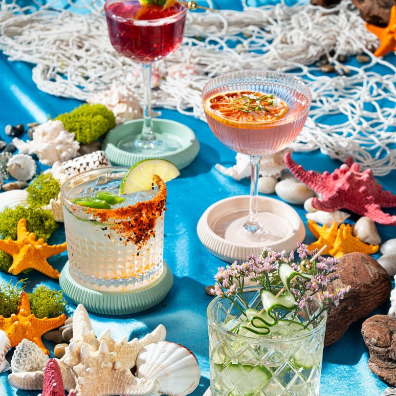 Four colorful cocktails on a blue surface, surrounded by seashells, starfish, and fishing net.