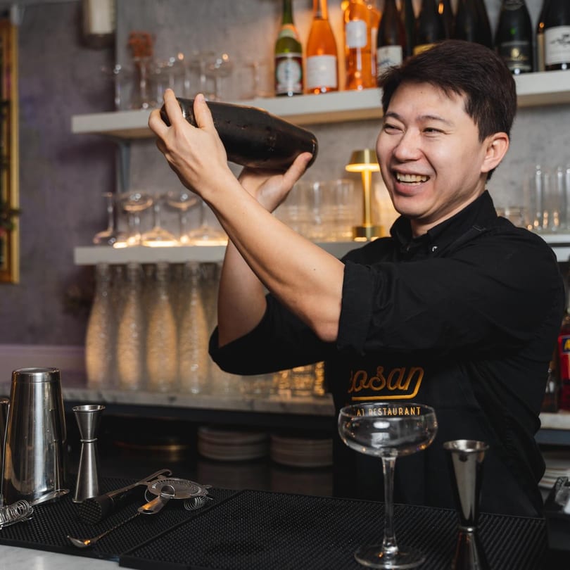 A smiling Khaosan bartender in a black apron vigorously shakes a cocktail shaker behind the bar.